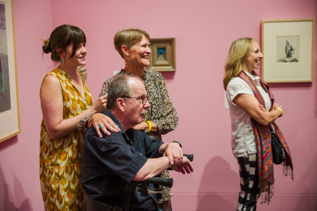 Four people gather in a pink gallery with framed artwork on the walls, they are facing a person speaking out of frame